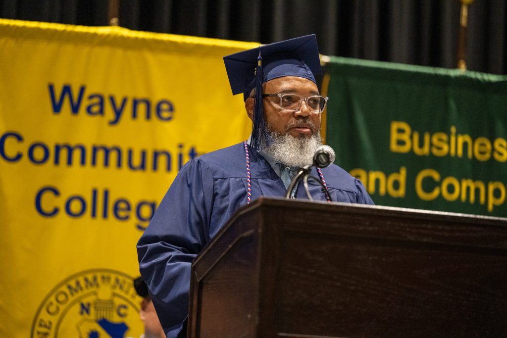 A man wearing a graduation cap and gown speaks at a podium.