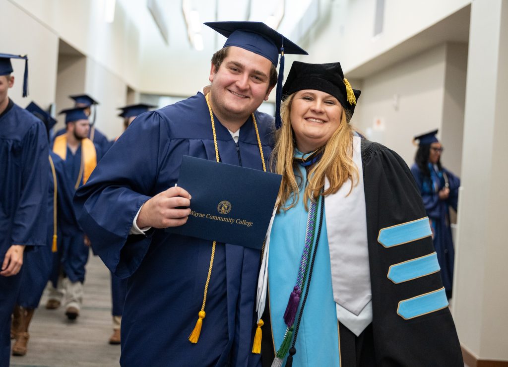 A man wearing a graduation cap and gown holds a diploma while standing next to a woman wearing a graduation cap and gown.
