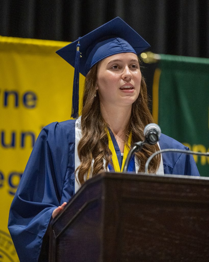 A girl wearing a graduation cap and gown speaks at a podium.