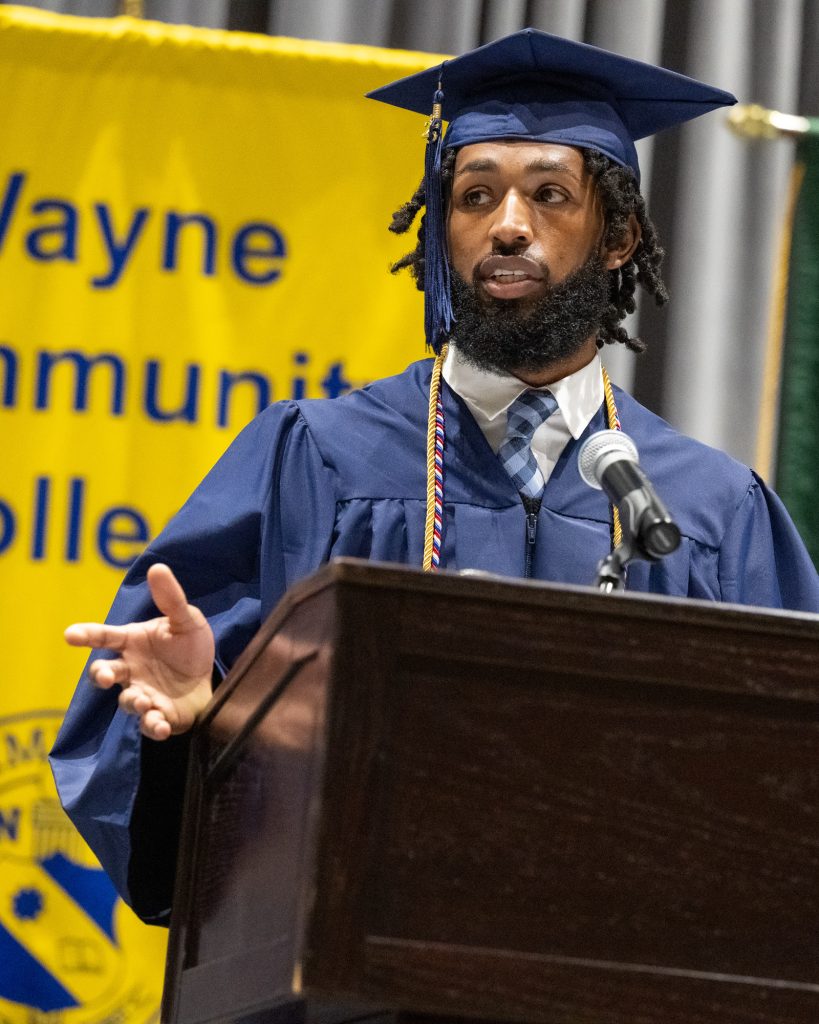 A man wearing a graduation cap and gown speaks while standing at a podium.