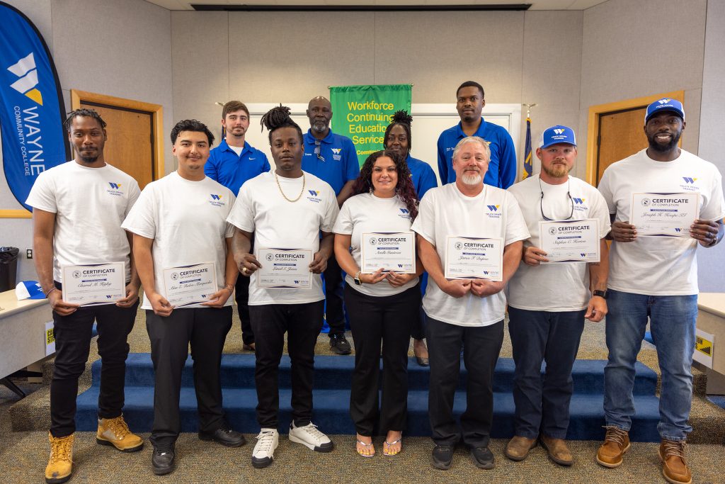 Individuals wearing white T-shirts hold certificates and stand in front of individuals wearing blue shirts.
