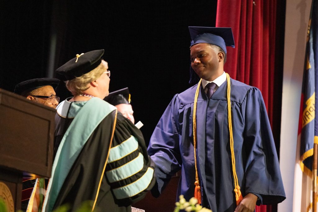 A man wearing a graduation cap and gown shakes the hand of a woman wearing a graduation cap and gown.