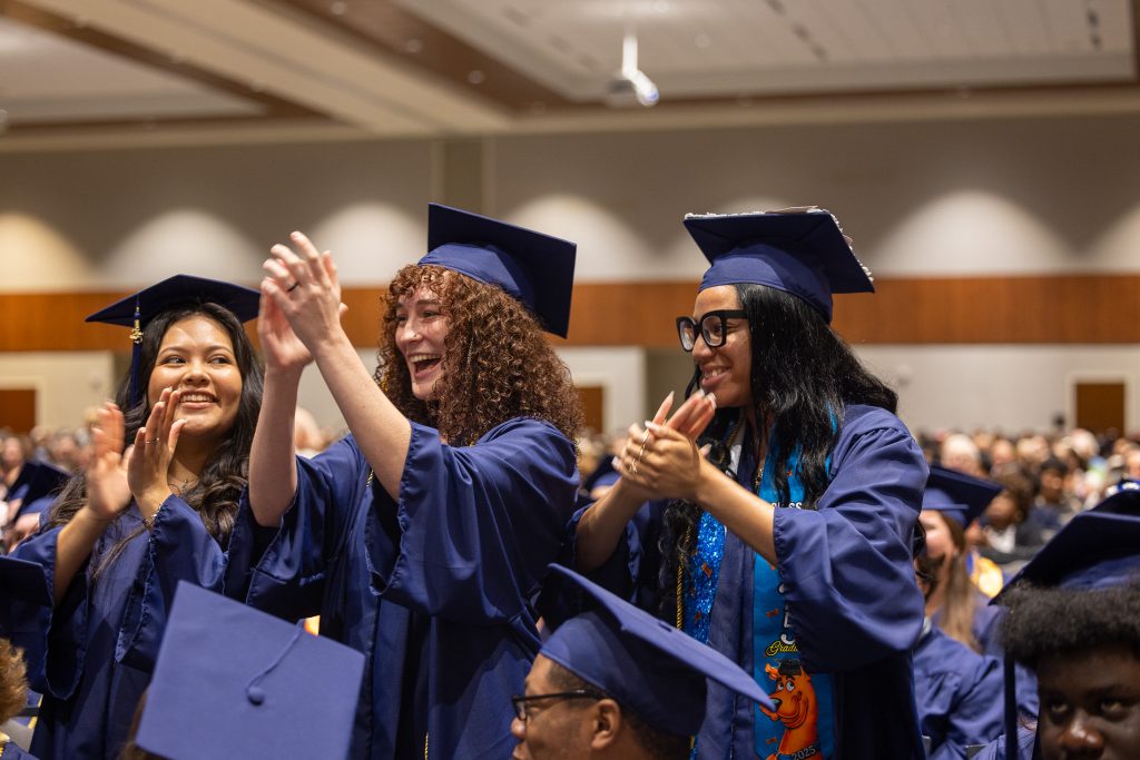 Three individuals wearing graduation caps and gowns stand and clap.