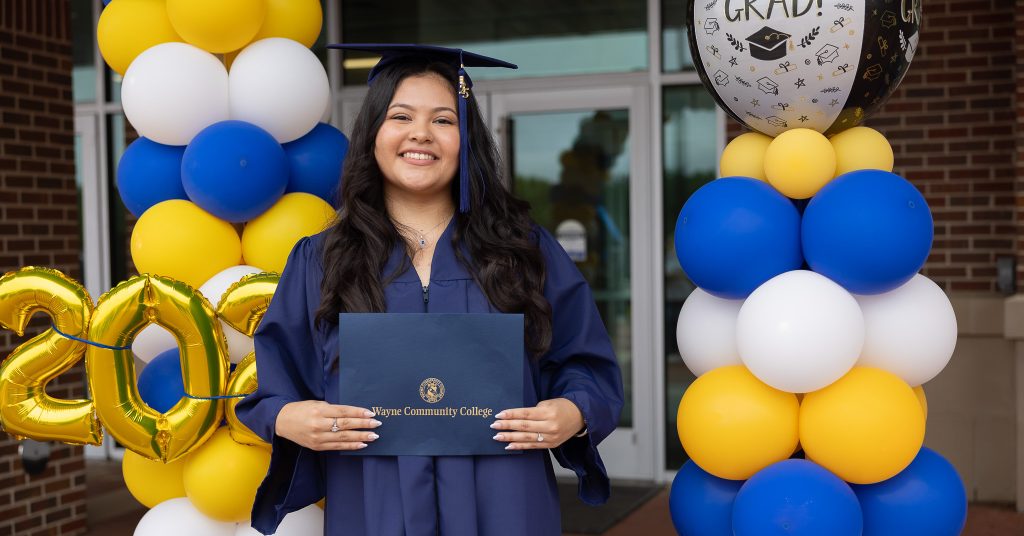 A woman stands in front of balloons while holding a diploma and wearing a graduation cap and gown.