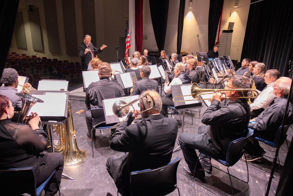 Individuals sit and play instruments while a man stands to direct them.
