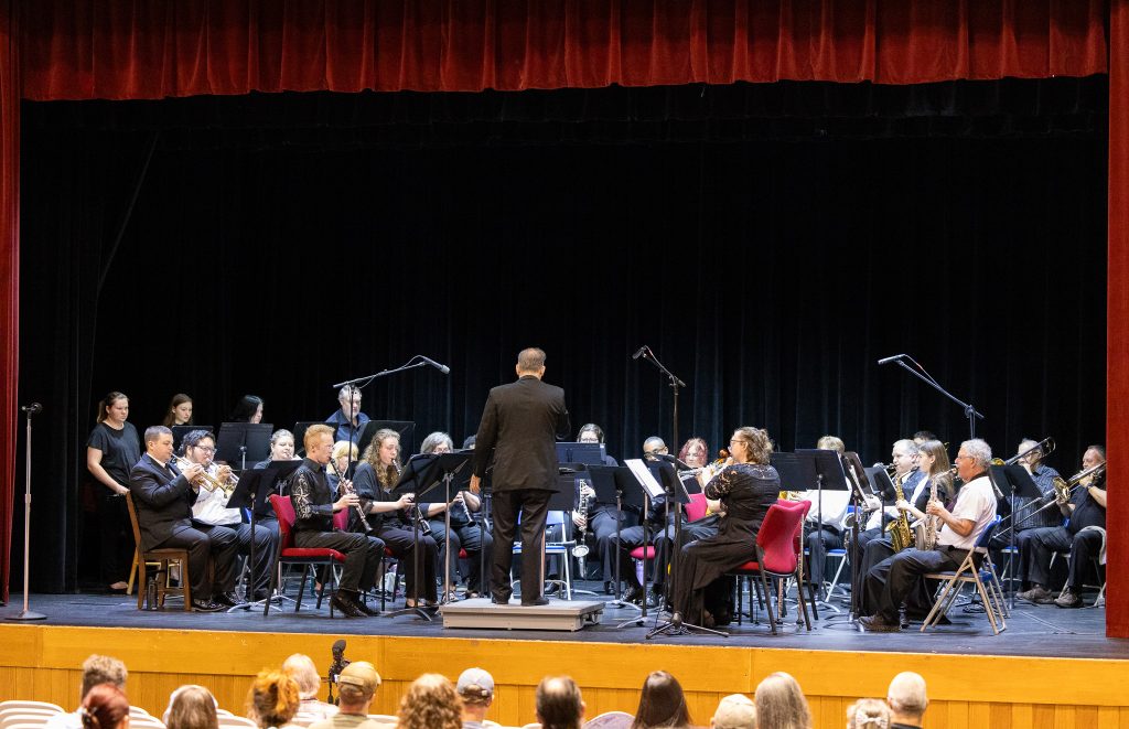 Musicians play their instruments on a stage while a conductor stands in front of them.