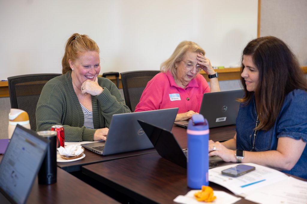 Three women sit at tables typing on laptops.