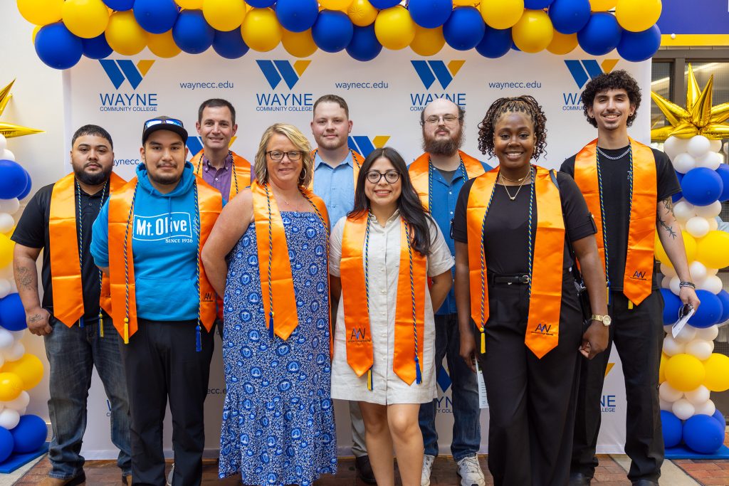 Individuals wearing orange graduation stoles stand in front of a backdrop with blue and yellow balloons.