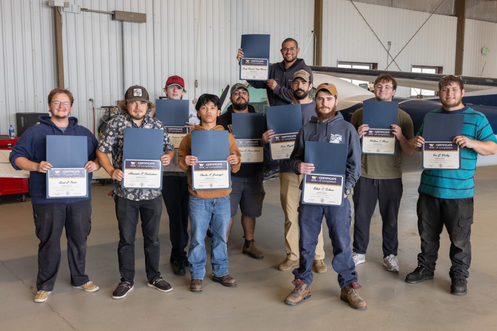 Students stand together in an airplane hangar while holding certificates.