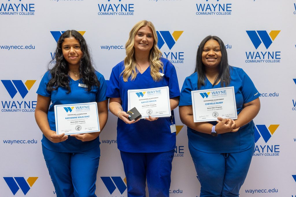 Three women wearing blue medical scrubs stand together holding certificates.