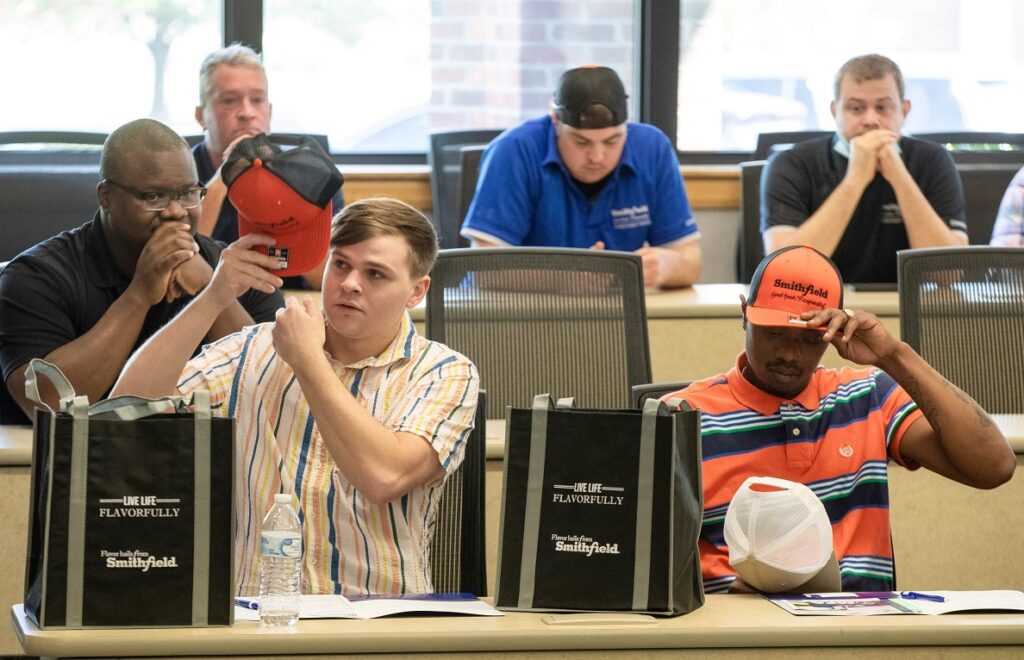 Two men putting on hats with company logos while men behind them watch