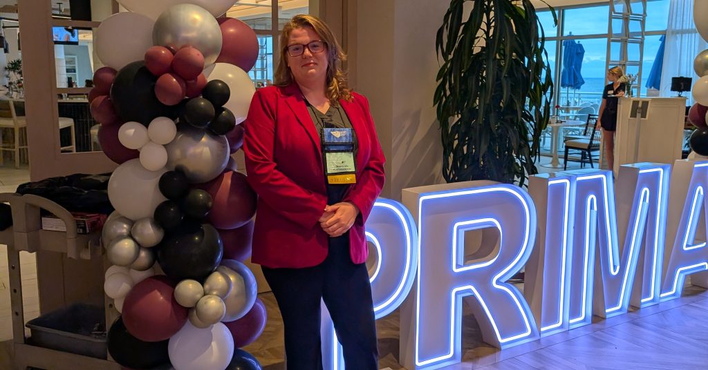 A woman in a red jacket stands in front of a sign and balloons.