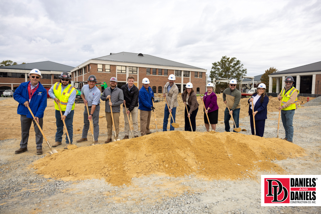 Individuals shovel a pile of dirt while wearing hard hats.