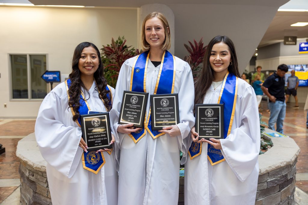 Three women stand together wearing graduation gowns while holding plaques.