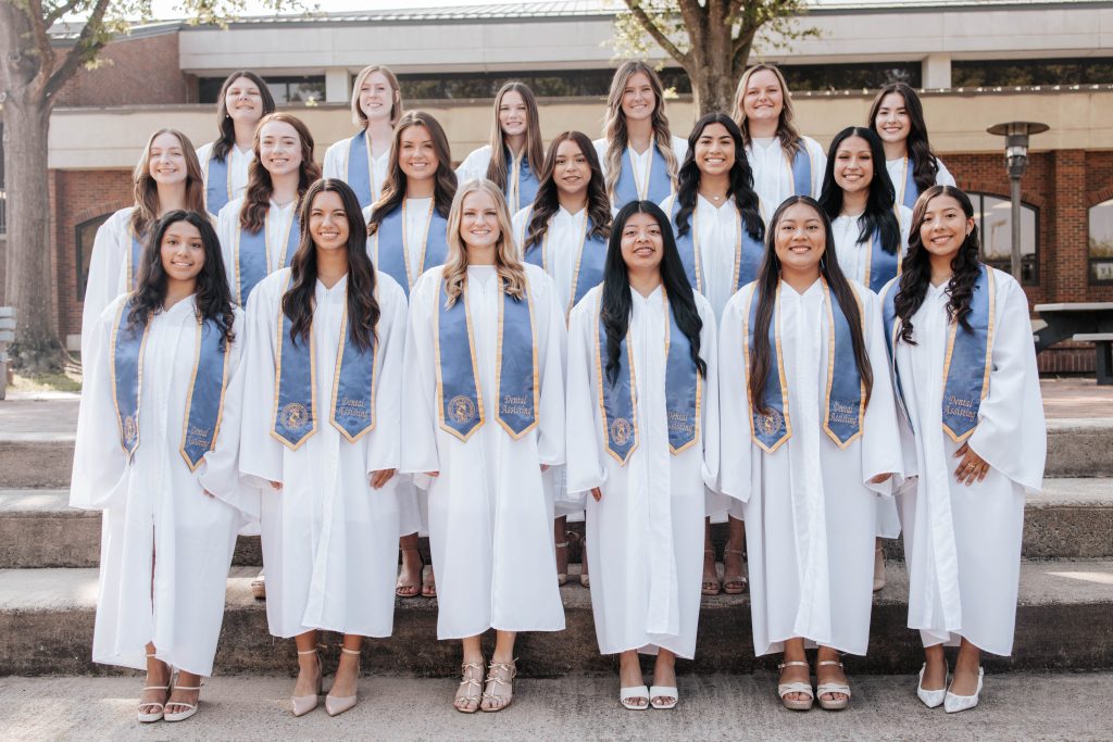 Individuals stand together wearing white graduation gowns and blue stoles.
