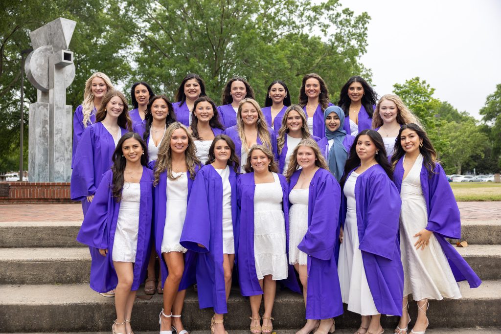 Graduates wearing purple graduation gowns stand together.