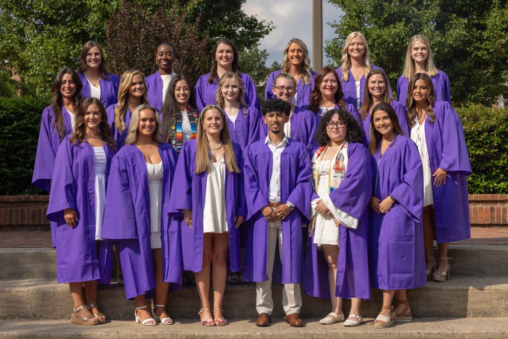 Students wearing purple graduation gowns stand together.