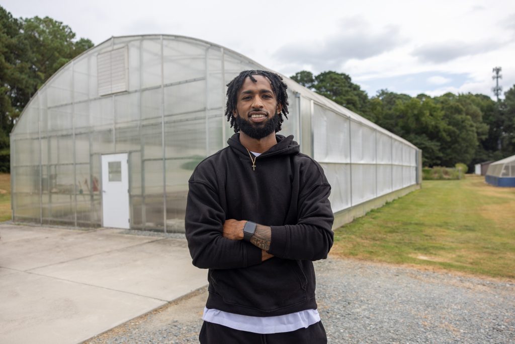 A man stands with his arms crossed in front of a greenhouse.