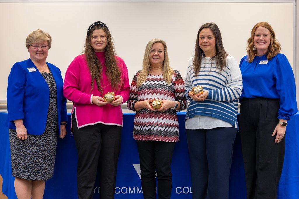 A group of women stand together while three of them hold golden apples.