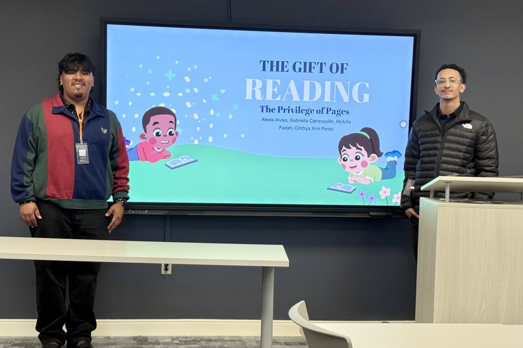 Two individuals stand beside a screen displaying children reading.