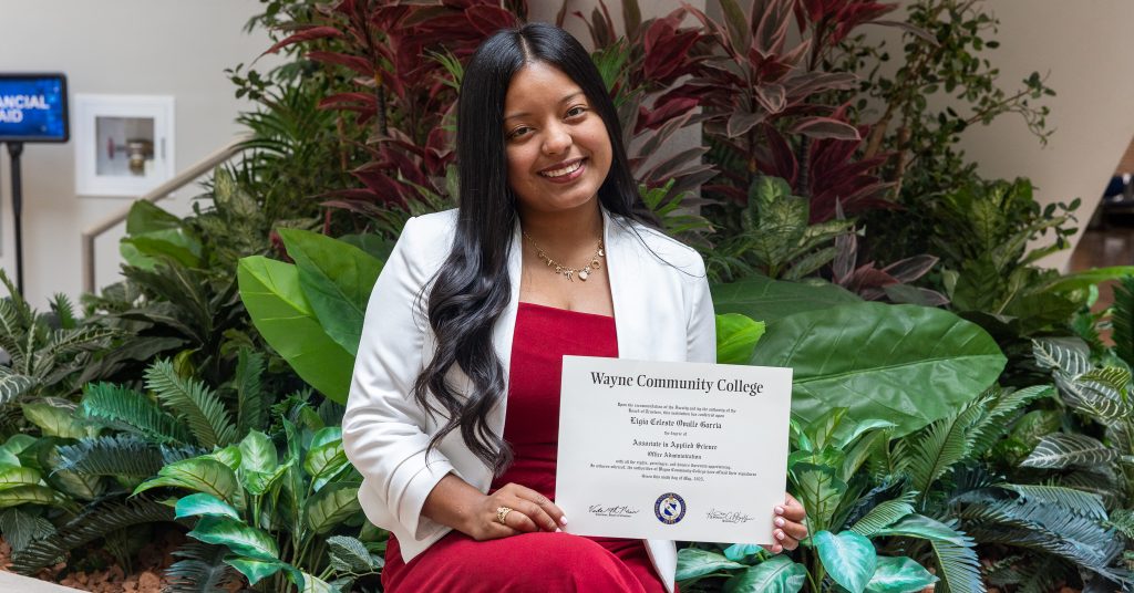 A woman sits in front of plants while holding a diploma.