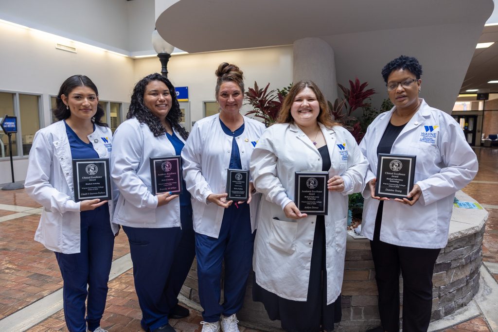 Five women wearing medical scrubs and lab coats hold plaques.