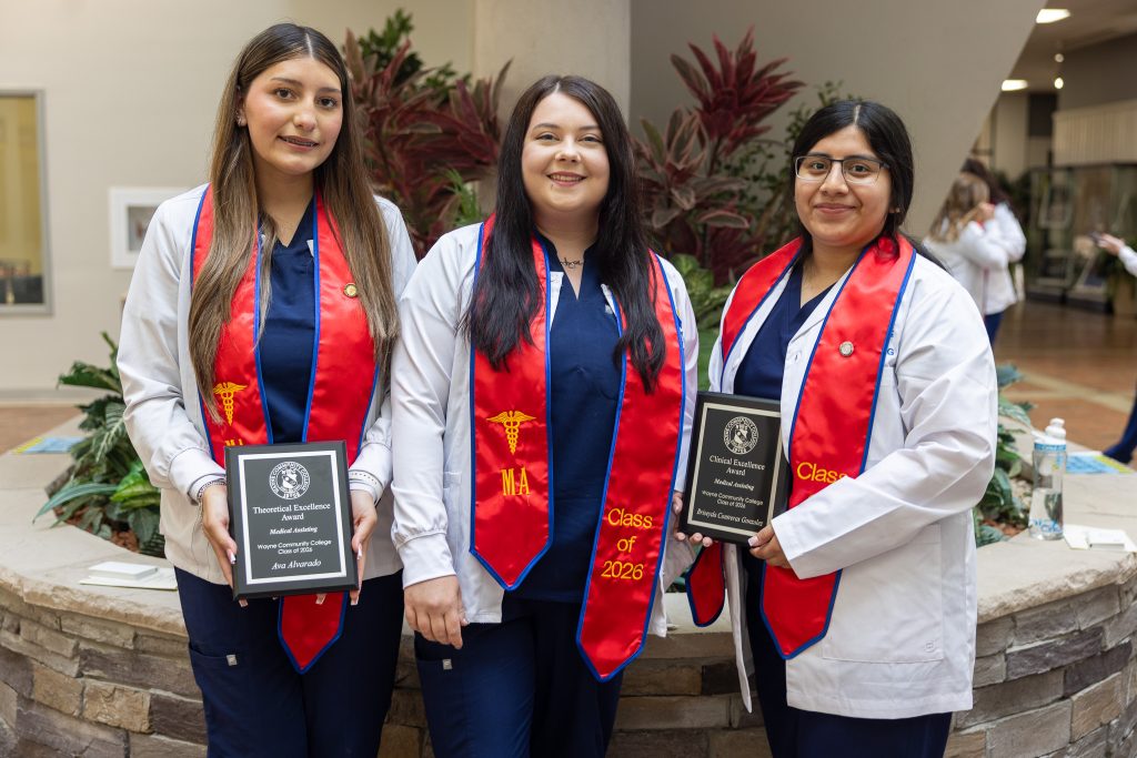 Students wearing white lab coats and red graduation stoles stand together holding plaques.