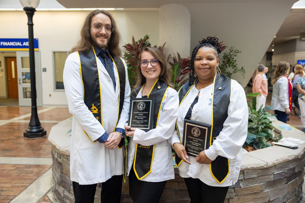 Students wearing lab coats and wearing black graduation stoles stand together holding plaques.