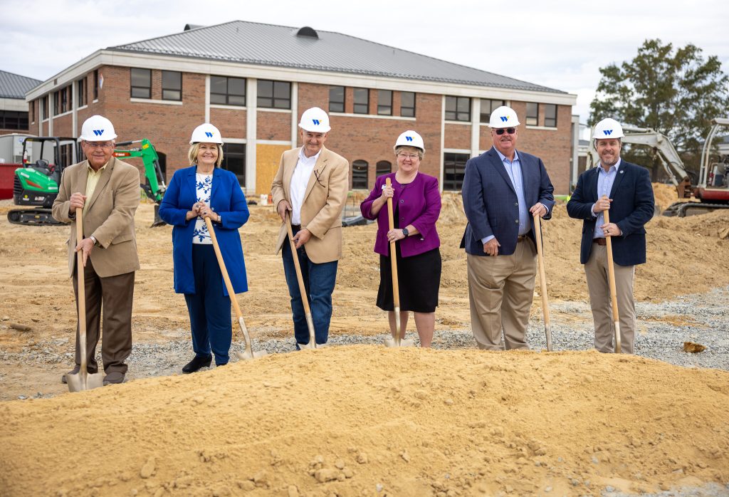 Individuals wearing hard hats stand in front of dirt while holding shovels.