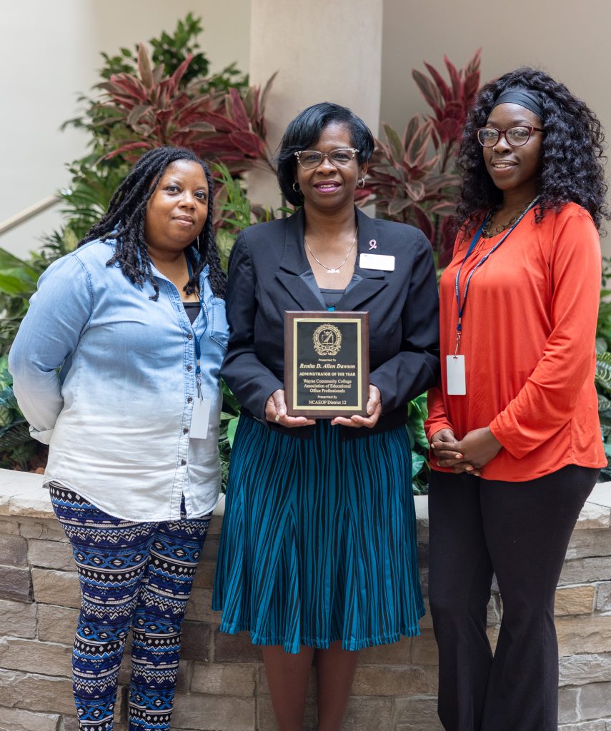 Three women stand together with the one in the middle holding a plaque.