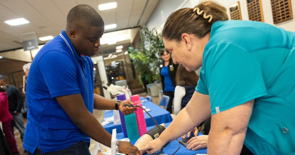 A woman wearing scrubs shows a young man how to take blood pressure using a mannequin's arm.