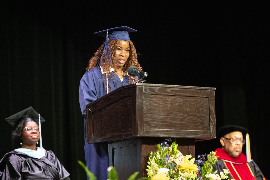 A woman wearing a graduation cap and gown speaks at a podium.
