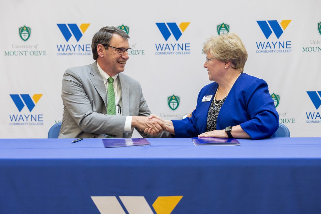 A man and woman sit behind a table, shaking hands.