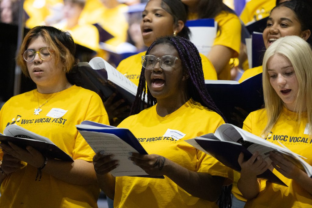 Students wear matching yellow shirts and hold sheet music while singing together.