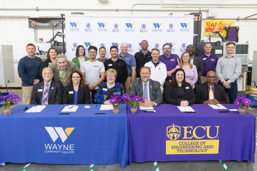Individuals stand and sit around two tables with blue and purple tablecloths.