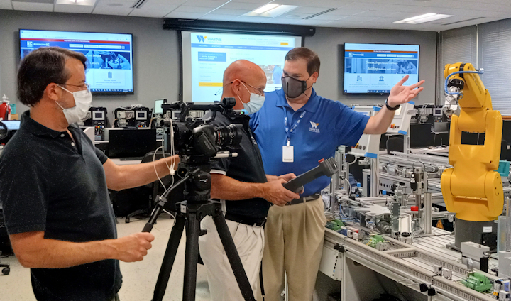 A camera ;man films two men gesturing in the middle of machinery.