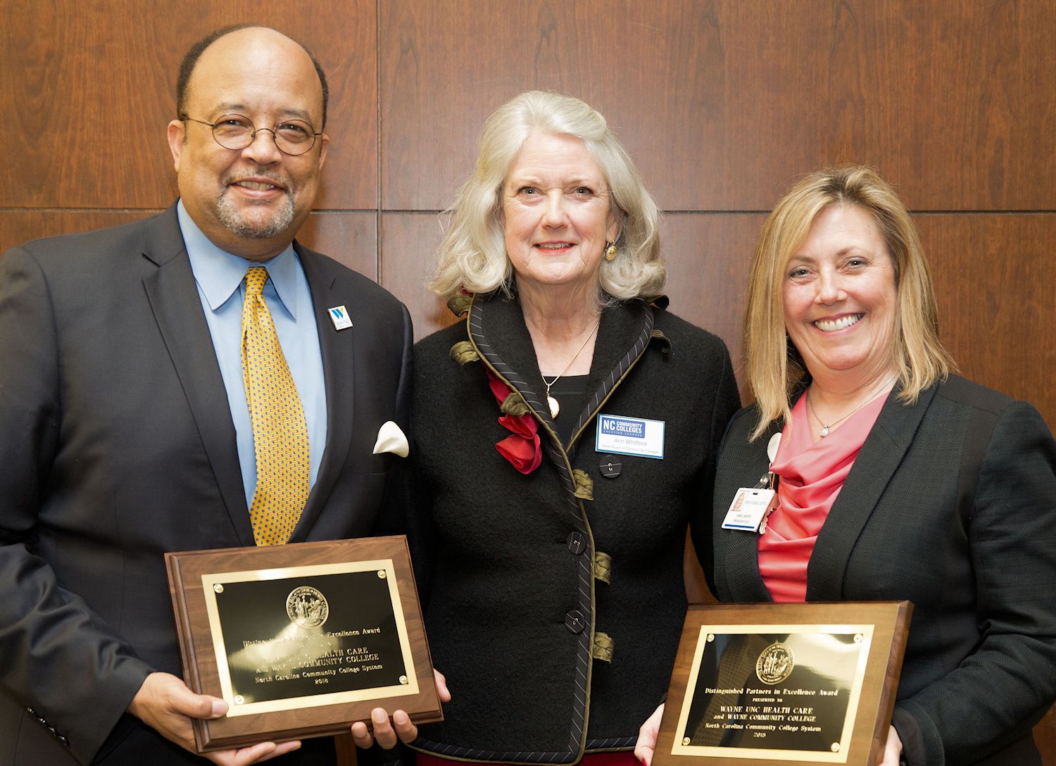 State Board of Community Colleges member standing with WCC President and Wayne UNC Health Care President and CEO, both of which have .Distinguished Partners in Excellence Award plaques in their hands.