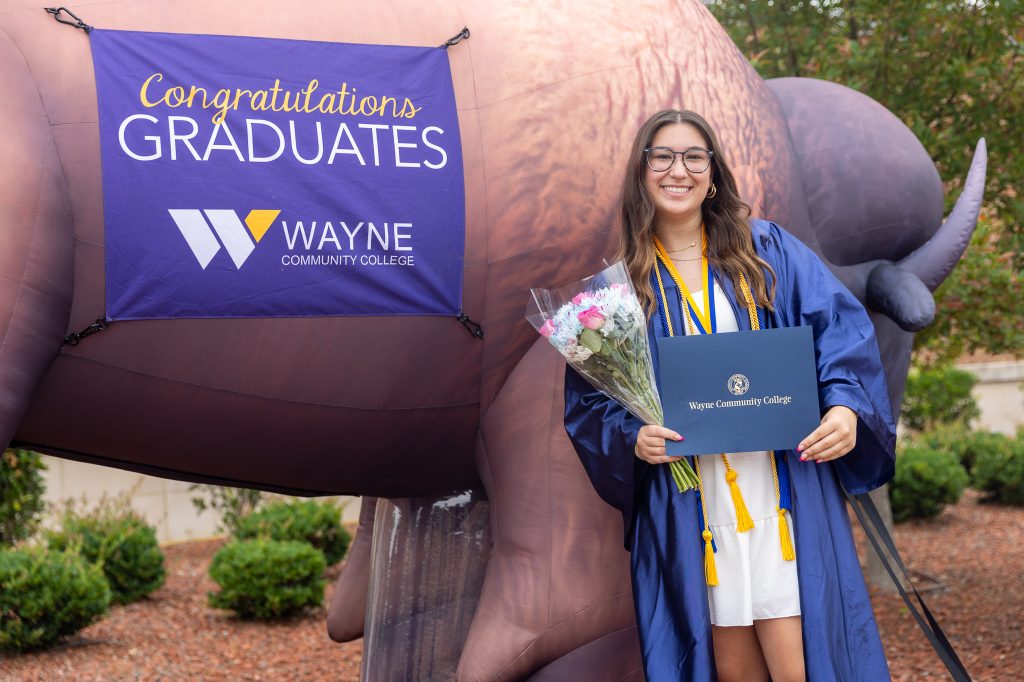 A student wearing a graduation gown holds a diploma and a bouquet in front of an inflatable bison.