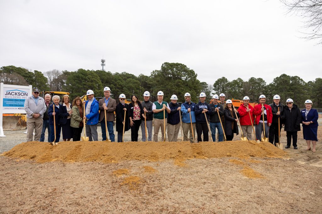 A group of people stand in front of a pile of dirt while wearing hard hats and holding shovels.