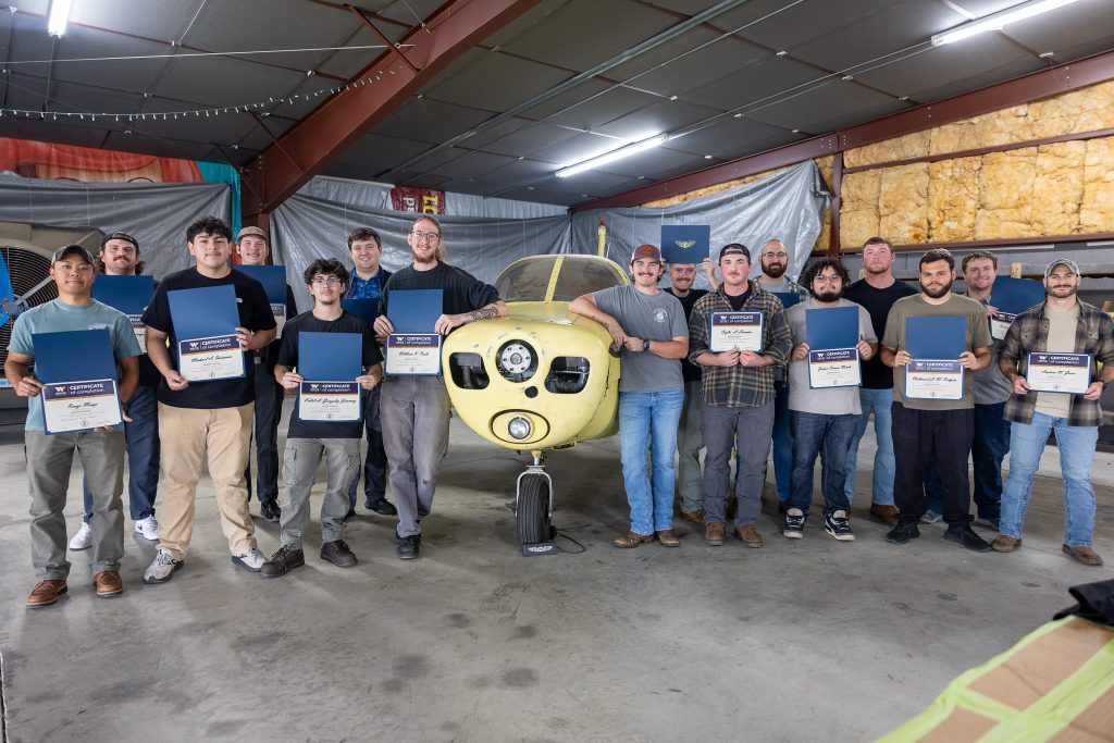 Students stand beside an airplane in a hangar while holding certificates.