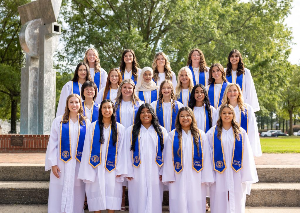 Individuals stand together wearing white graduation gowns and blue stoles.