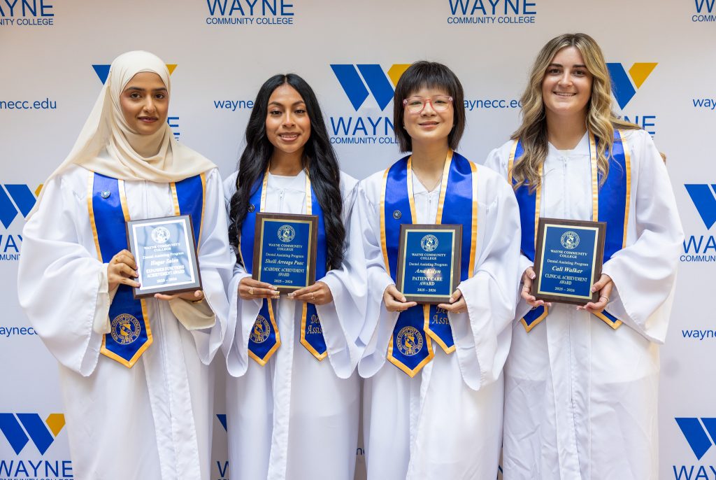Four women stand together in white graduation gowns holding plaques.
