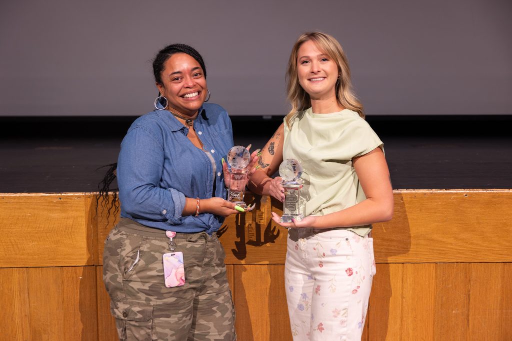 Two women stand beside each other holding trophies.