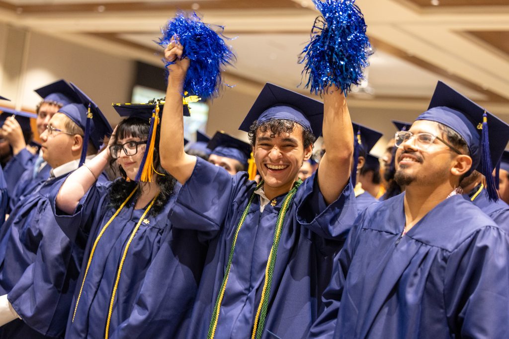 Graduates wearing caps and gowns stand together with one waving pom poms.