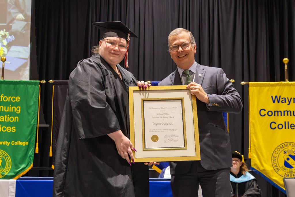 A woman wearing a graduation cap and gown and a man in a suit stand together holding a framed certificate.