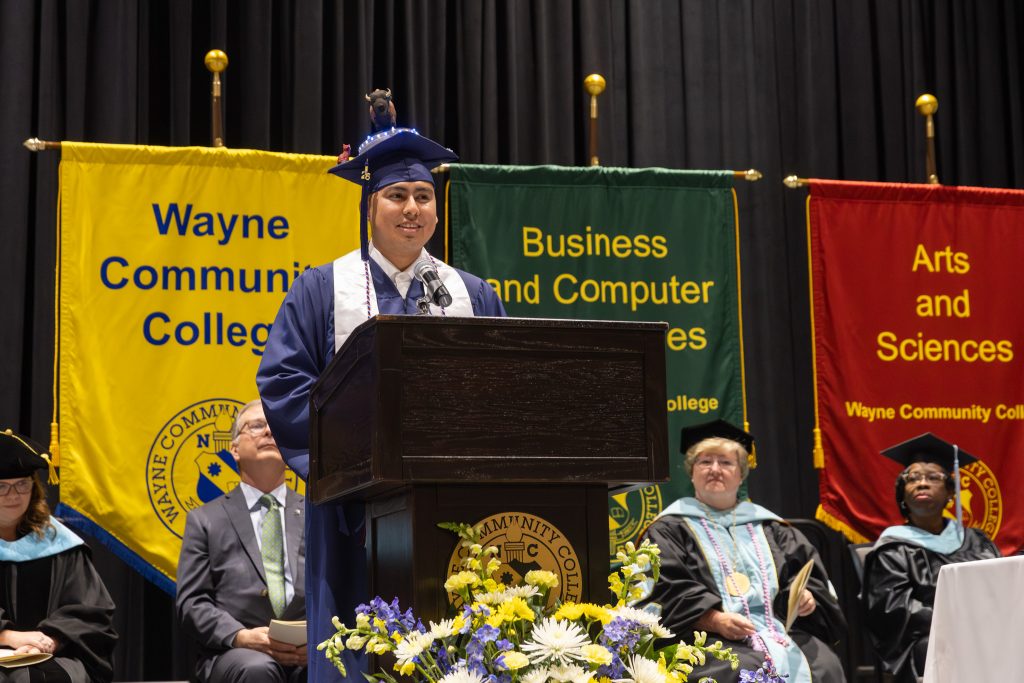 A man speaks at a podium while wearing a graduation cap and gown.