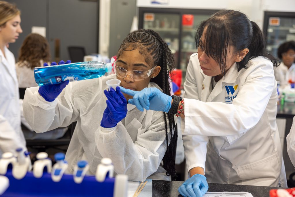 A girl pours liquid into a cup while another person assists.
