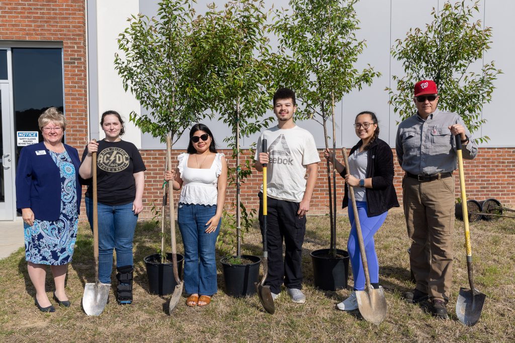 Individuals stand in front of trees while holding shovels.